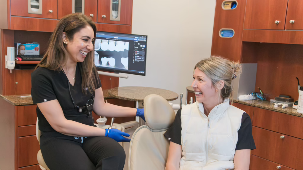 Dentist with blue gloves smiling at a patient in a dental chair, monitor showing dental X-rays in the background.