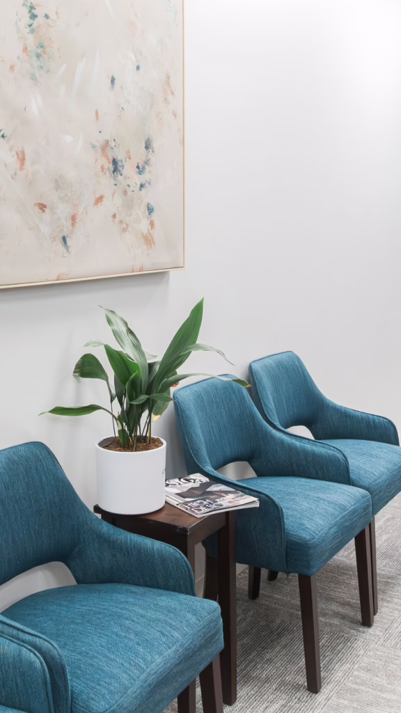 Waiting area with three teal upholstered chairs around a small side table holding a white pot plant and magazines, abstract painting on the wall above.