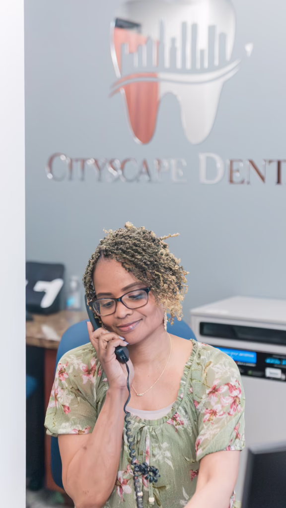 Receptionist with glasses talking on a corded phone at a dental clinic reception, Cityscape Dental logo in the background.