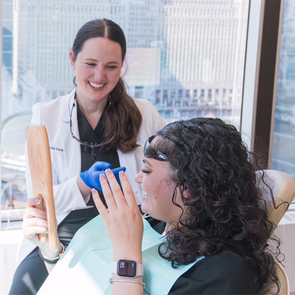 Dentist in a white coat and blue gloves smiles at a patient seated in a dental chair, holding a mirror for the examination.