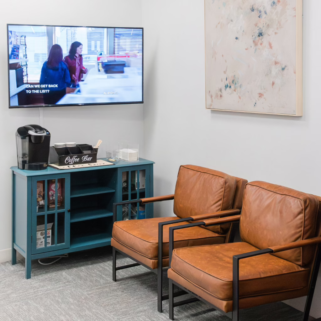 Teal coffee bar with a Keurig machine, cups, and snacks beside two brown leather armchairs in a small waiting area, with a wall-mounted TV and an abstract painting above.