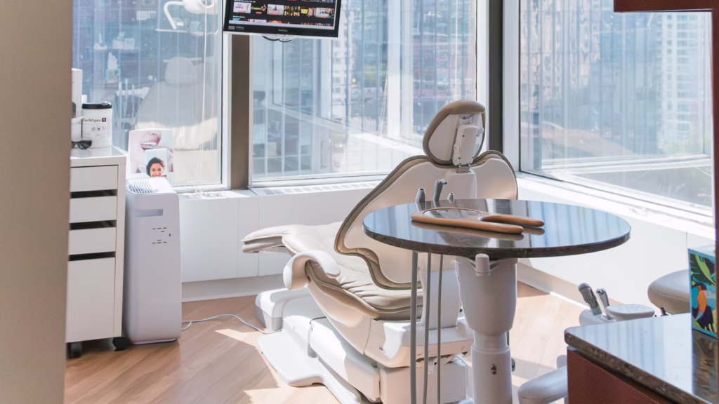 Modern dental chair setup in a bright clinic with large windows and a cityscape view; dental tools and a round table nearby.